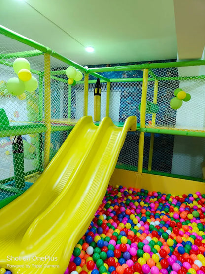 A bright indoor playground with a yellow double slide into a ball pit, decorated with balloons.