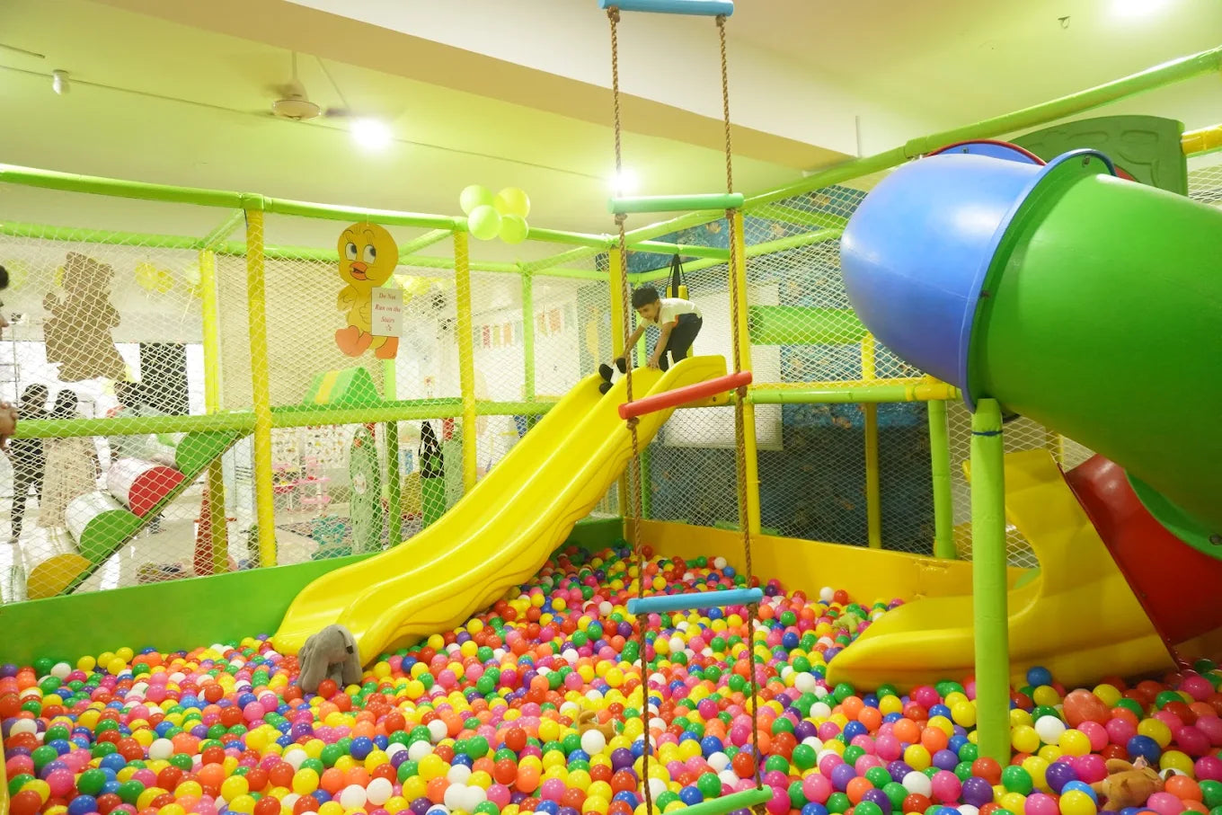 A child plays on a yellow slide in a colorful ball pit at an indoor playground.