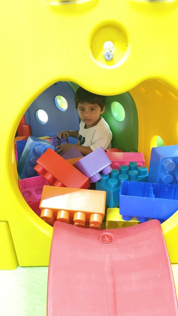 A young child plays with large building blocks inside a yellow play structure.