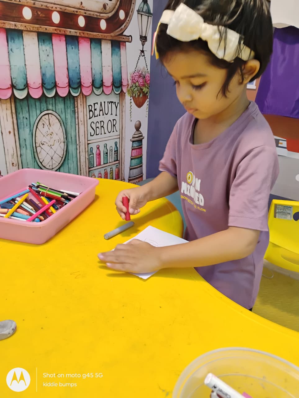 A young child draws on a yellow table with crayons, surrounded by art supplies.