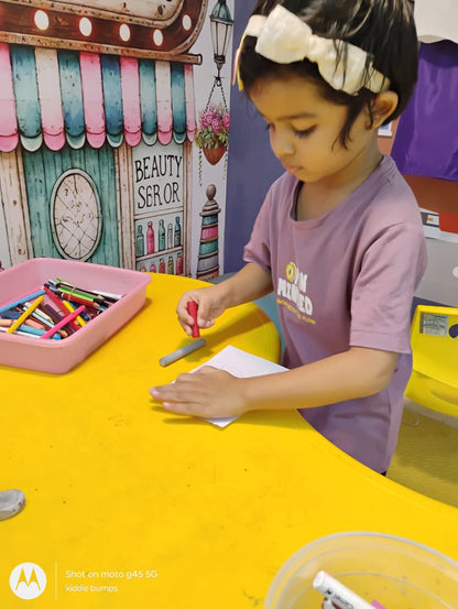 A young child draws on a yellow table with crayons, surrounded by art supplies.