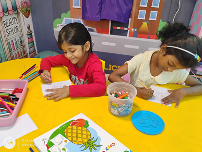 Two young girls are coloring at a yellow table, with art supplies around them.