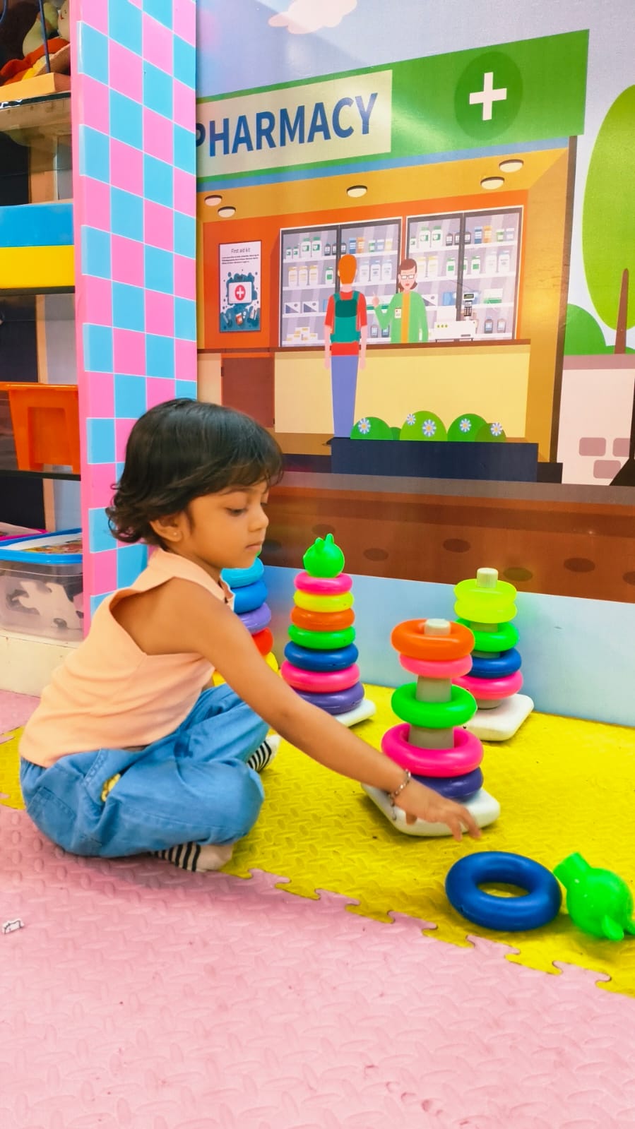 A young child plays with stacking rings in front of a colorful pharmacy-themed backdrop.