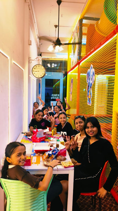 A group of young girls are gathered around a table with food and drinks, celebrating in a colorful indoor play area.