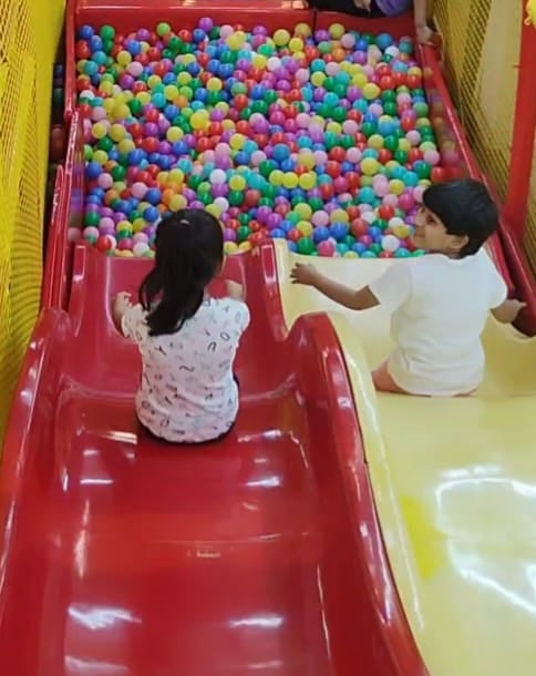 Two young children slide down a red slide into a ball pit filled with colorful balls.
