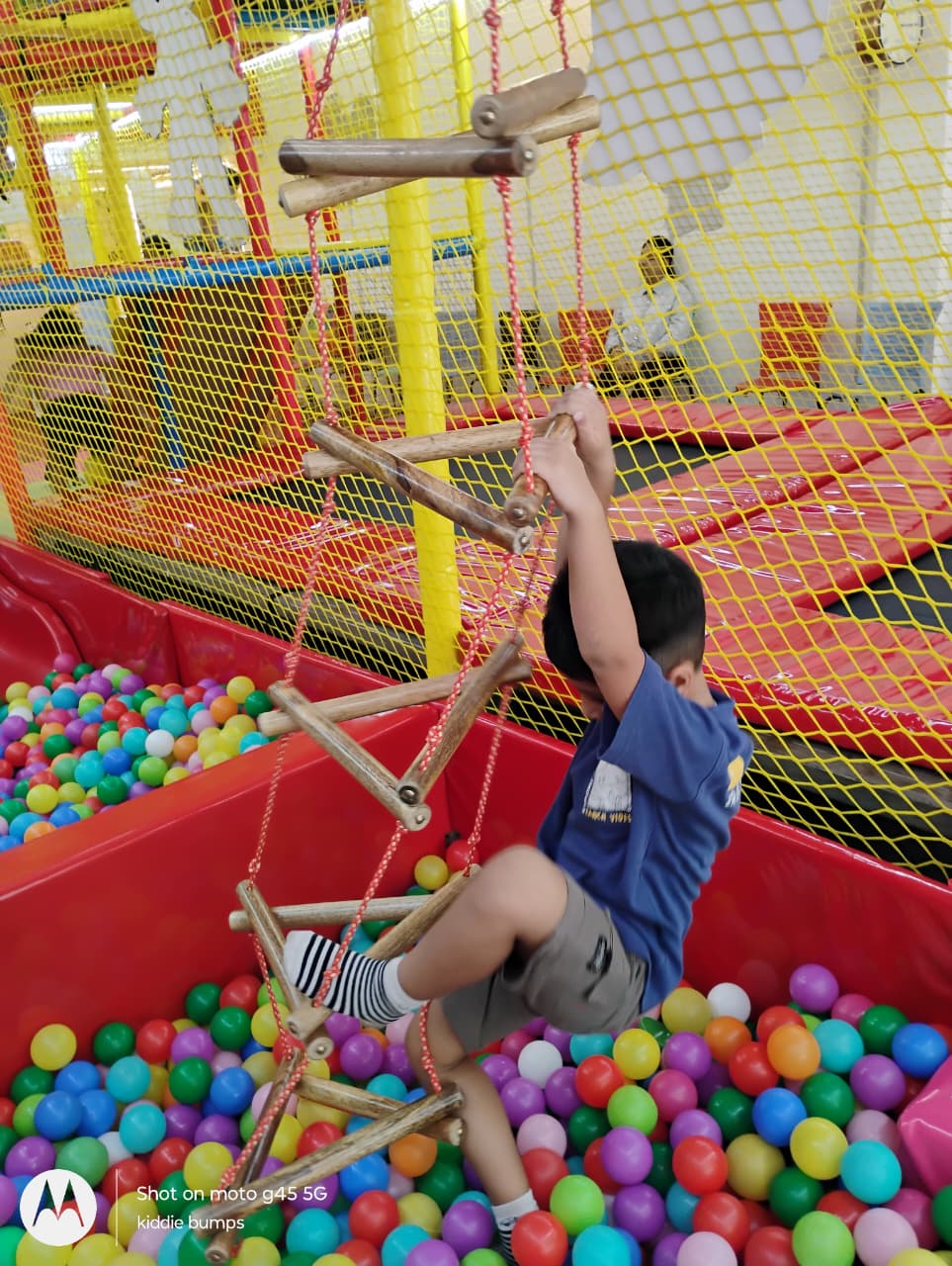 A young child climbs a rope ladder over a ball pit in a colorful indoor playground.