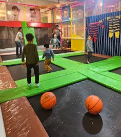 Children playing in an indoor trampoline park with colorful walls and basketballs on the floor.