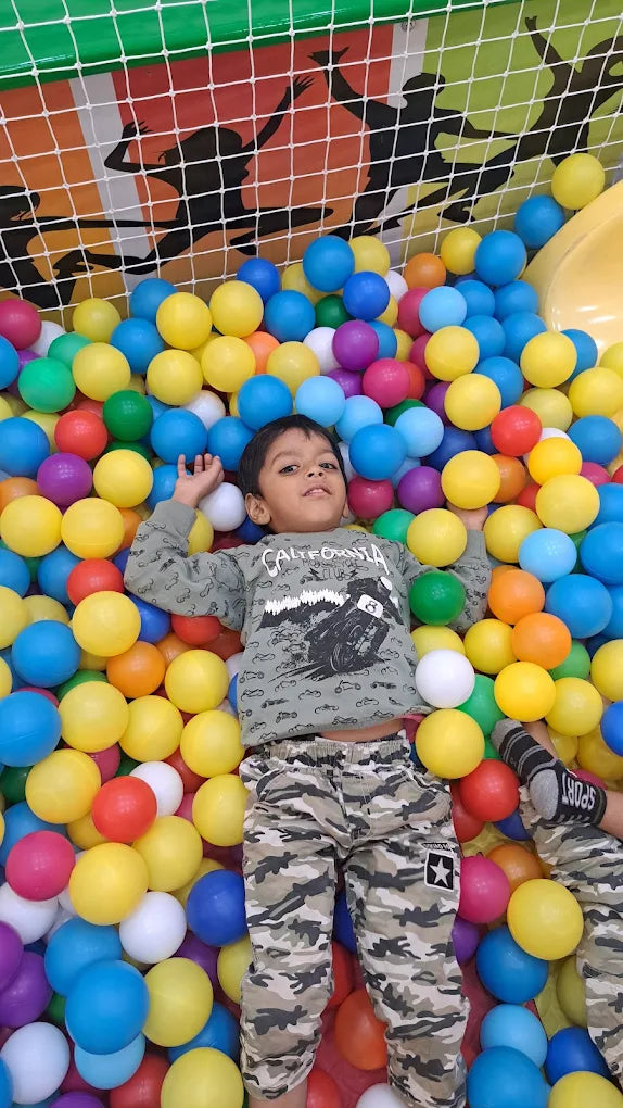 A young boy lies in a ball pit, surrounded by colorful balls. He is wearing a grey shirt and camouflage pants.