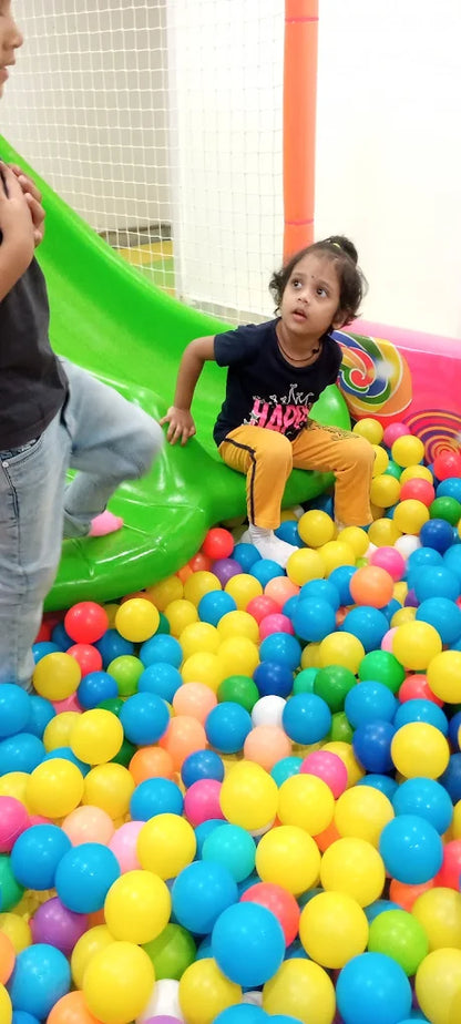 A young child sits in a ball pit next to a green slide, looking up.