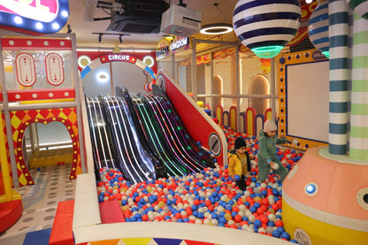 Children play in a colorful ball pit with slides in a circus-themed indoor playground.