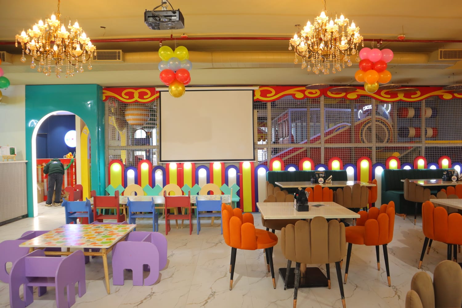 A colorful indoor playground and dining area for children, with tables, chairs, and a play structure.