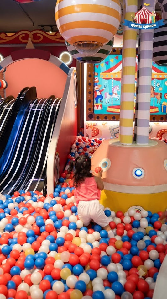A young child plays in a ball pit at an indoor playground. The ball pit is filled with red, blue, and white balls.