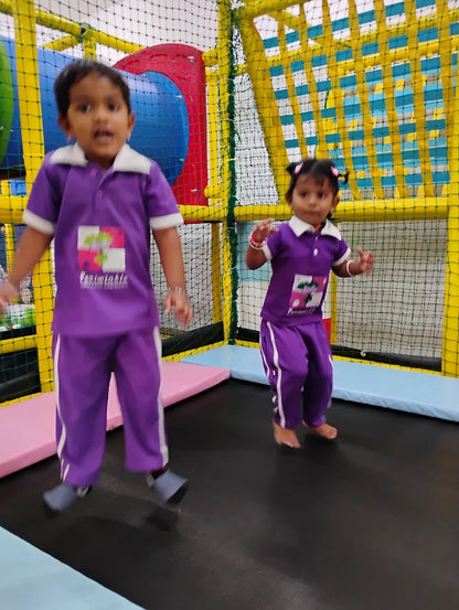 Two young children in purple jumpsuits are happily jumping on a trampoline in an indoor play area.