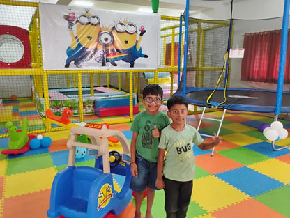 Two young boys stand in a colorful indoor playground with a trampoline and play structures.