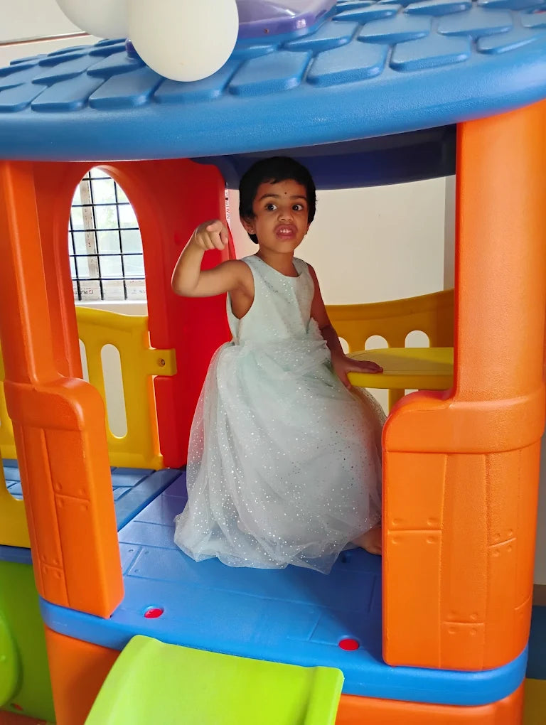 A young girl in a sparkly dress poses inside a colorful playhouse.