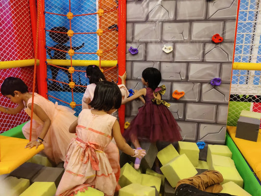 Young girls in dresses play in a colorful indoor playground with a climbing wall and soft blocks.