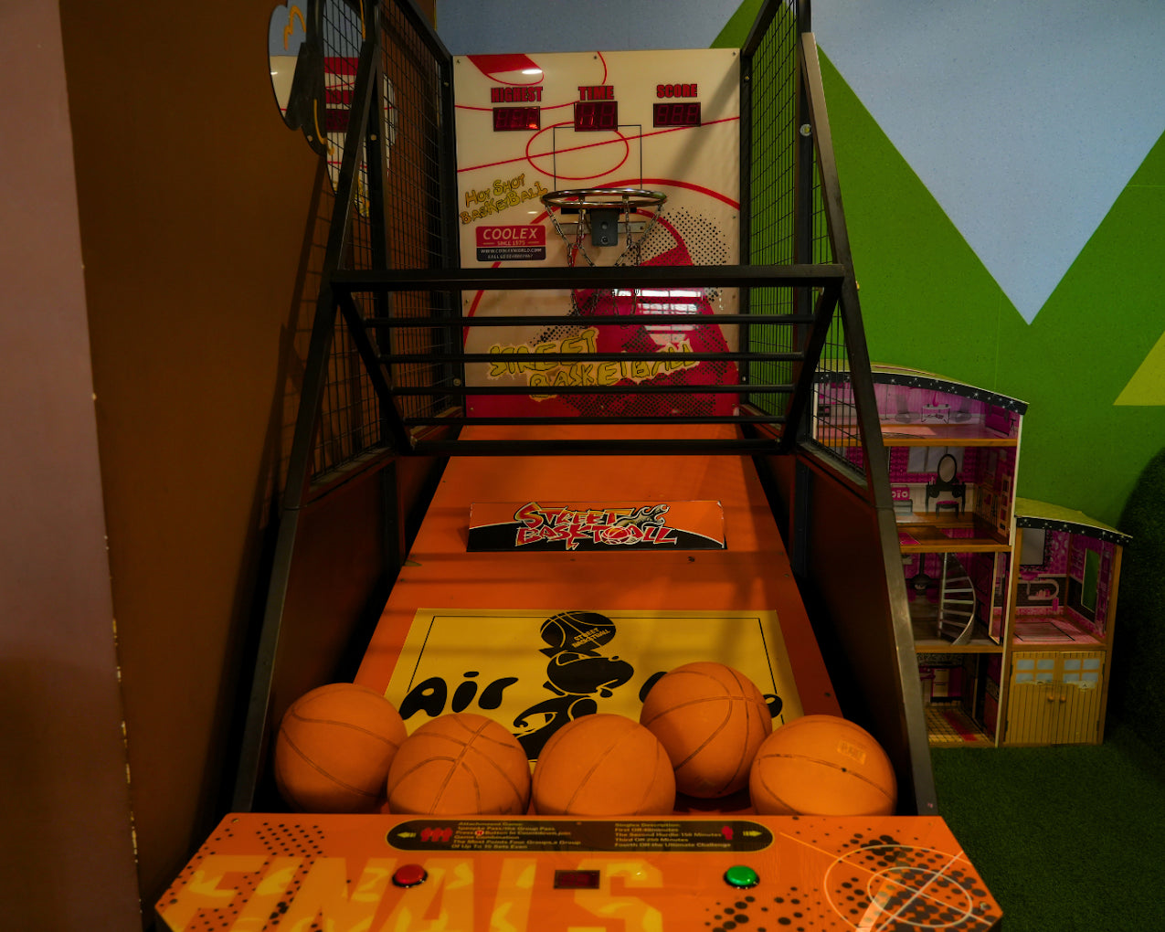 An arcade basketball game with orange balls and a dollhouse in the background.