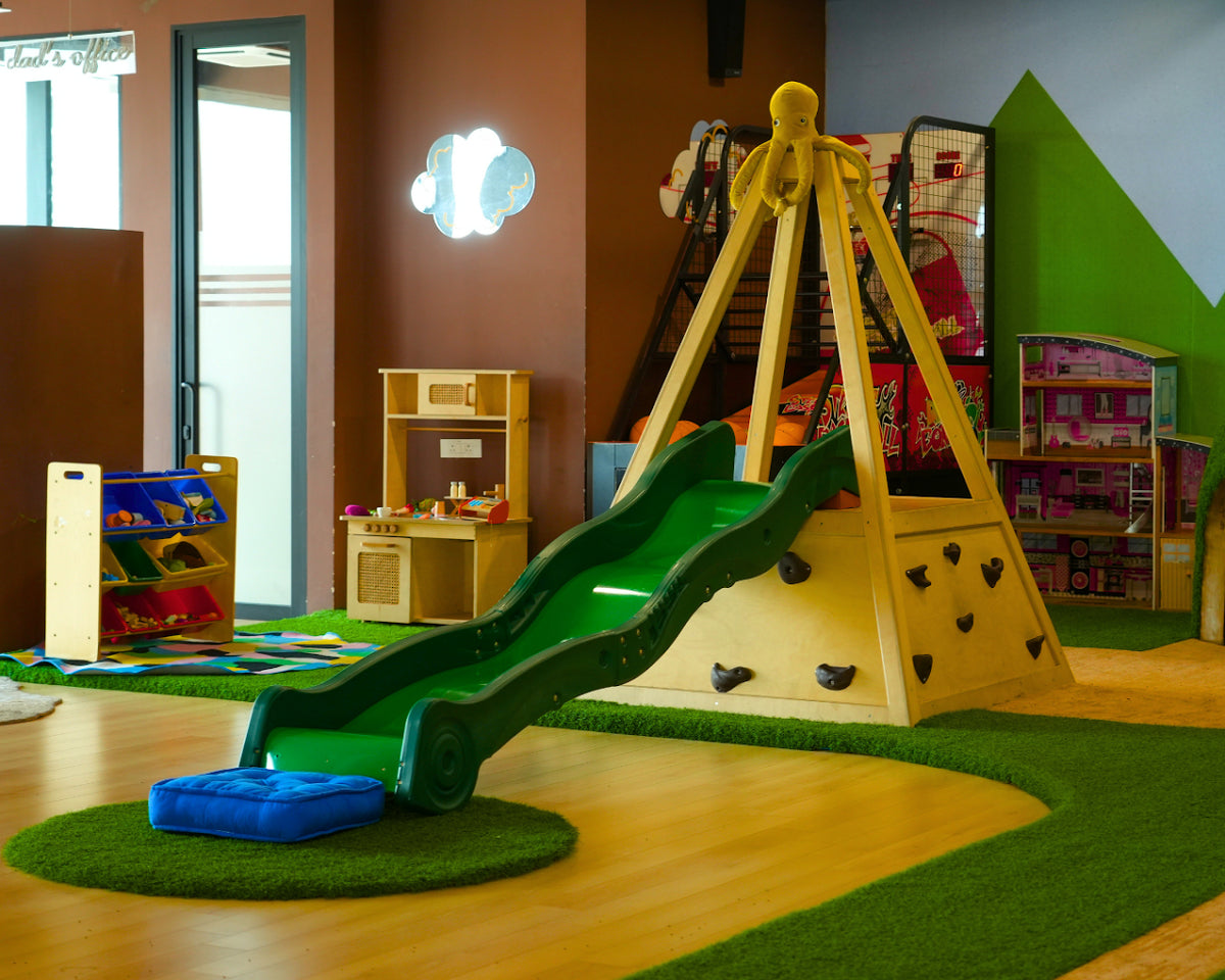 A colorful indoor playground with a slide, climbing wall, and toy kitchen for young children.