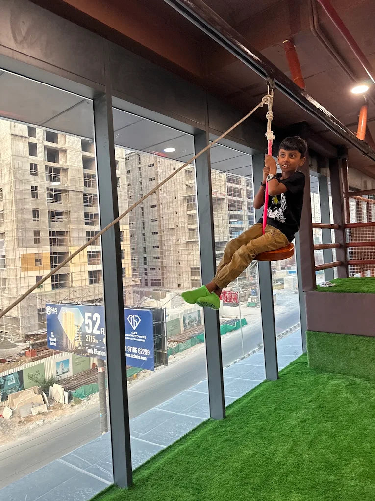A young boy rides a zipline indoors, with a city construction site visible through large windows.