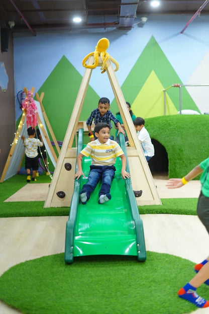 A young boy slides down a green slide at an indoor playground, while other children play nearby.