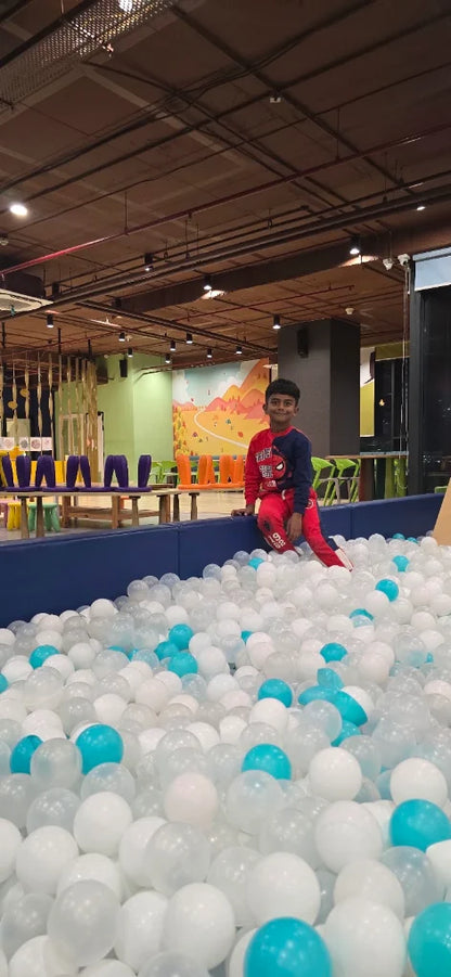 A young boy in a Spider-Man outfit sits on the edge of a ball pit filled with white and blue balls. He is in an indoor play area with colorful decorations.