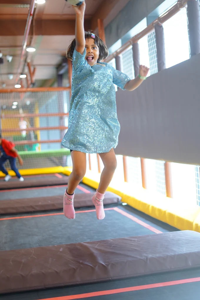 A young girl in a sparkly blue dress jumps high on a trampoline.