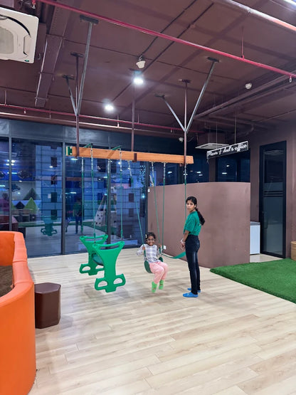 A young girl swings on a green swing while a teenager watches in a play area.