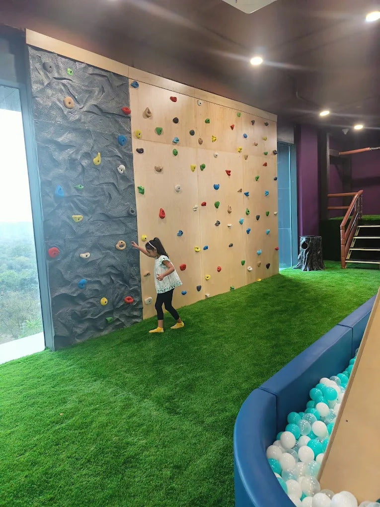 A young girl climbs a colorful rock wall in a play area with artificial grass and a ball pit.