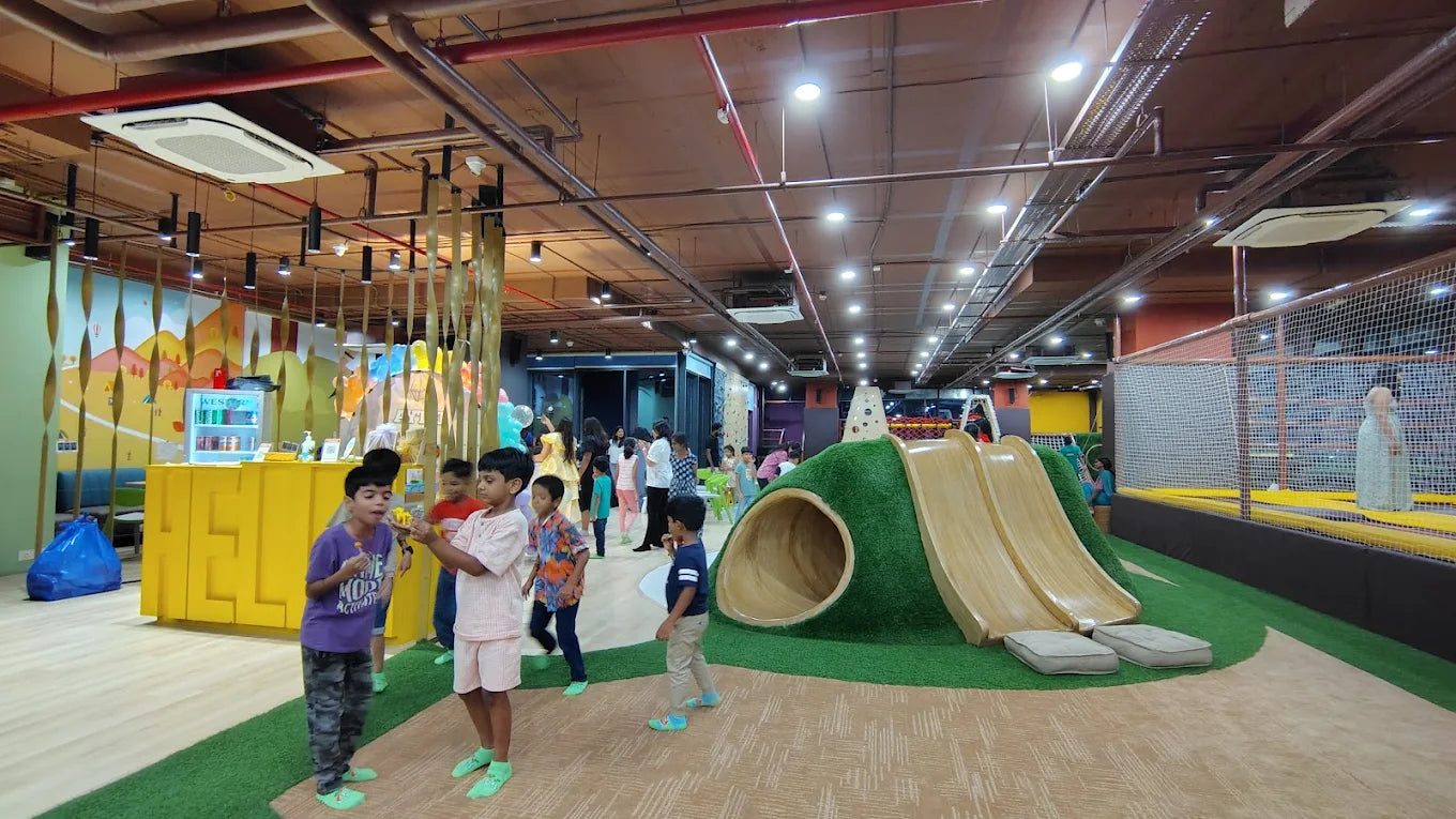 Children play in a colorful indoor playground with slides and a trampoline area.