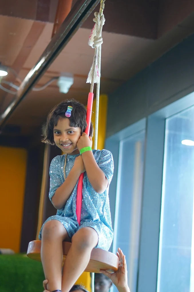 A young girl in a sparkly blue outfit smiles while sitting on a swing. She holds onto the swing's rope, which is attached to a ceiling beam.