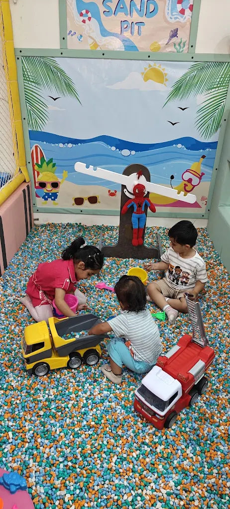 Young children play with toy trucks in a colorful ball pit with a beach-themed backdrop.
