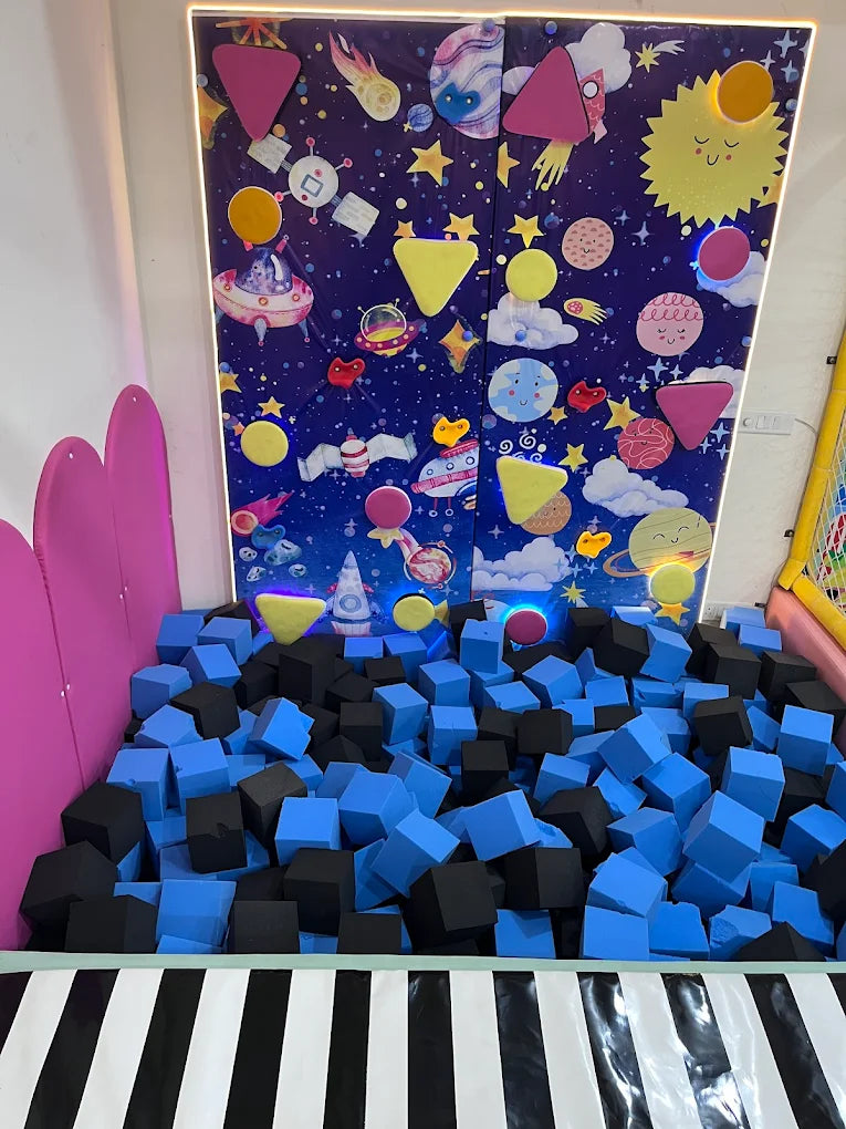 A ball pit filled with blue and black foam cubes, with a space-themed mural on the wall.
