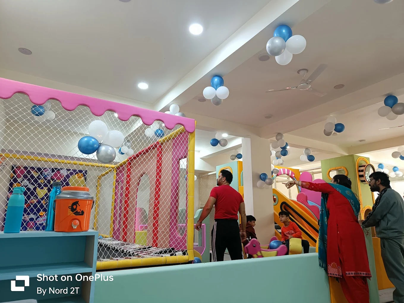 Adults and children are in a brightly colored indoor play area decorated with balloons.