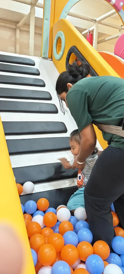 A woman helps a young child climb stairs in a ball pit.