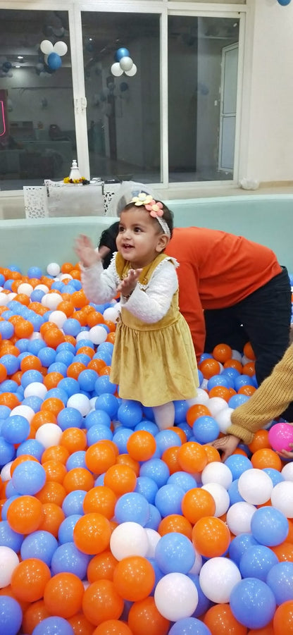 A baby girl in a yellow dress claps her hands in a ball pit filled with orange, blue, and white balls.