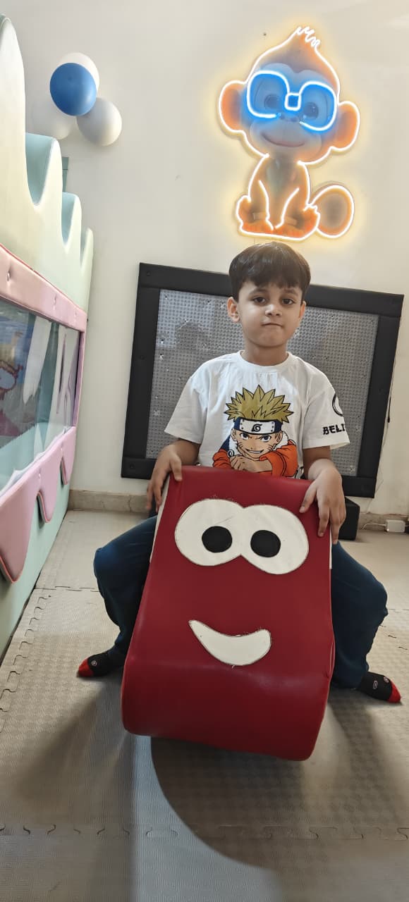 A young boy sits on a red, smiling cushion in a playroom with a neon monkey sign.