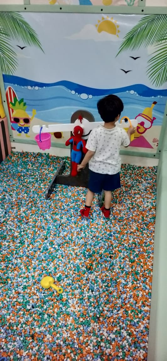 A young child plays in a ball pit with a Spiderman toy and a yellow shovel. The ball pit has a beach-themed backdrop.