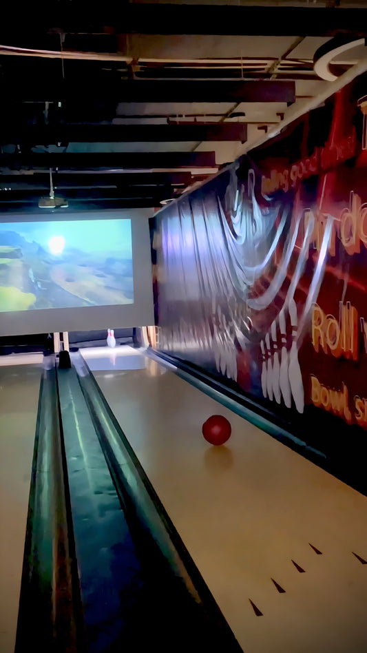 A red bowling ball rests on a lane in front of a projector screen showing a landscape.