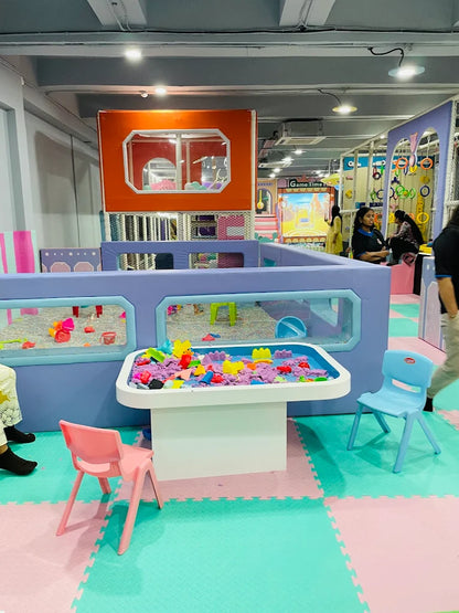 Children play in a colorful indoor playground with a sand table and soft play structures.