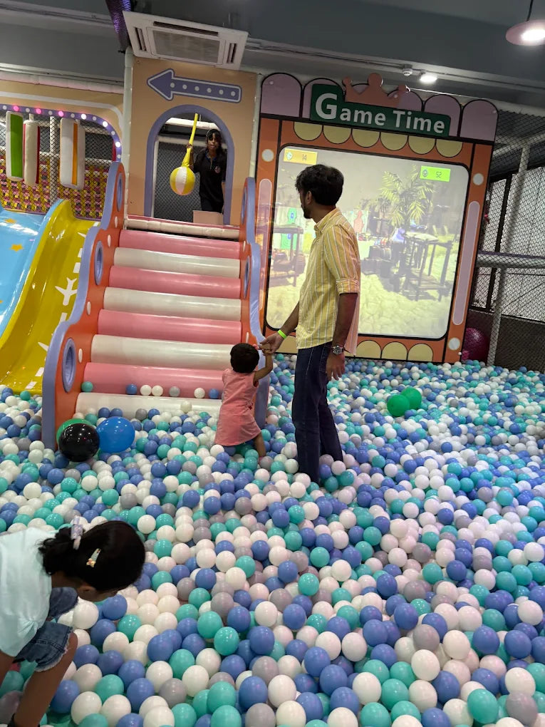 A man watches a child play in a ball pit at an indoor playground.