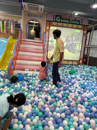 A man watches a child play in a ball pit at an indoor playground.