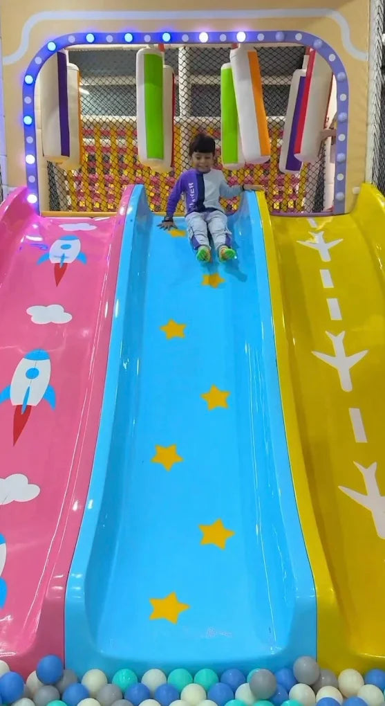 A young child slides down a blue slide in a colorful indoor playground. The slide is decorated with yellow stars.