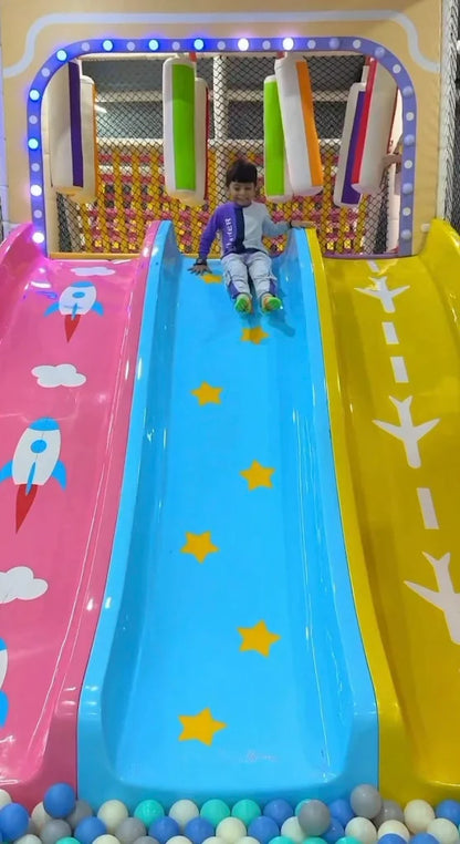 A young child slides down a blue slide in a colorful indoor playground. The slide is decorated with yellow stars.