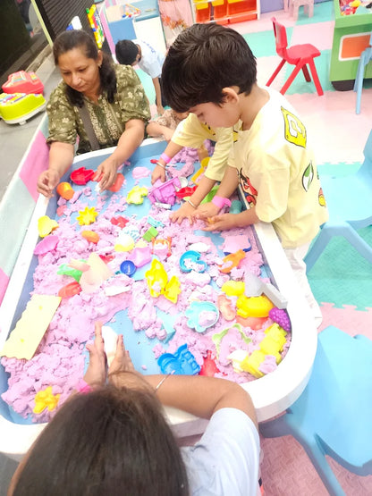 Children play with kinetic sand and toys at a sensory table, supervised by an adult.