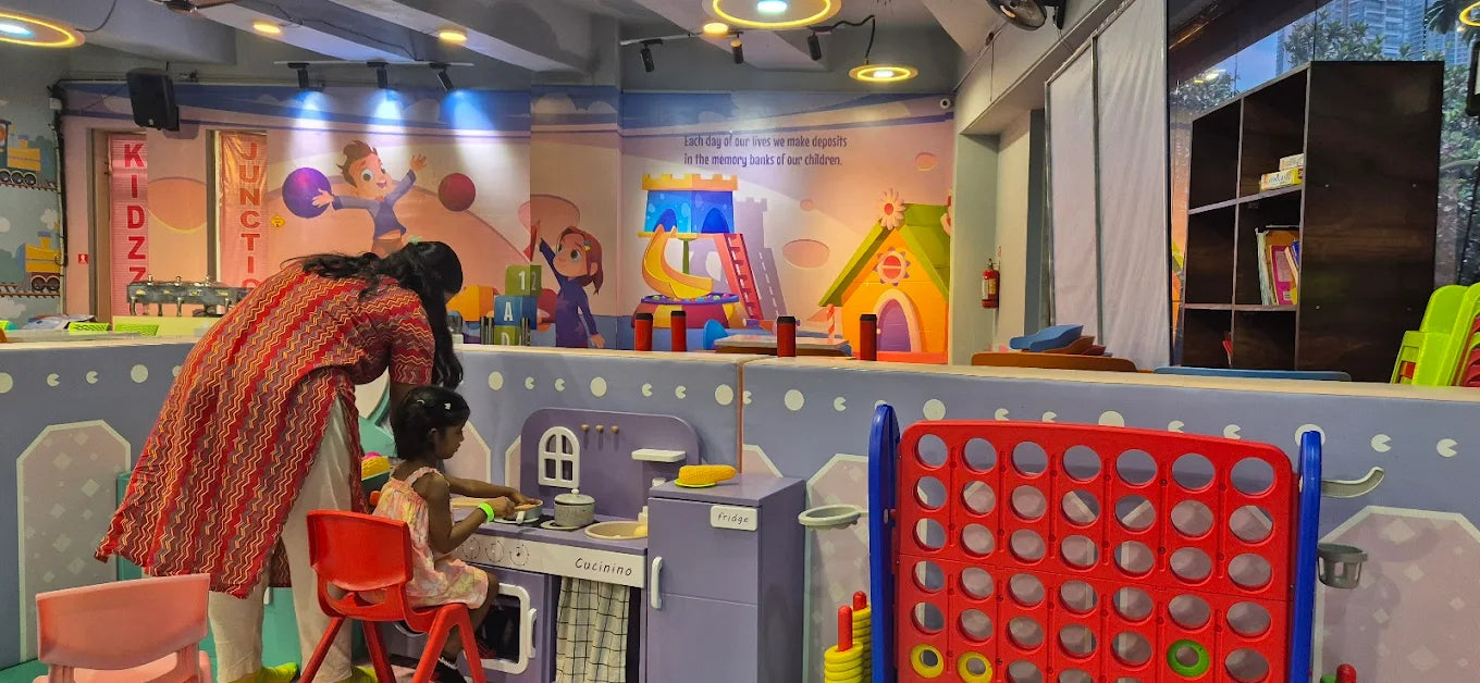A woman and two young children play in a colorful indoor play area with a toy kitchen and giant connect four.