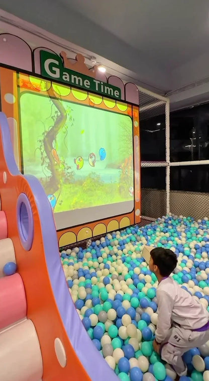 A young boy plays a projected game in a ball pit. The game screen shows cartoon birds in a forest setting.