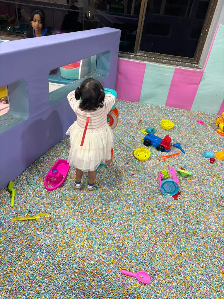 A young child plays in a ball pit with colorful toys, while an older child watches from behind a purple structure.