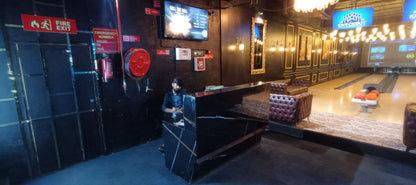 A man sits at a reception desk in a bowling alley with lanes visible in the background. Fire exit and emergency number signs are posted on the wall.