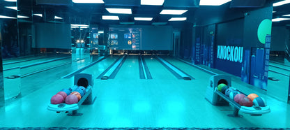 A bowling alley with blue lighting, showing bowling balls and lanes. A screen displays various images.
