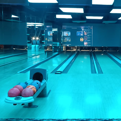 A bowling alley with multiple lanes, a ball return rack with colorful balls, and a large screen displaying videos.